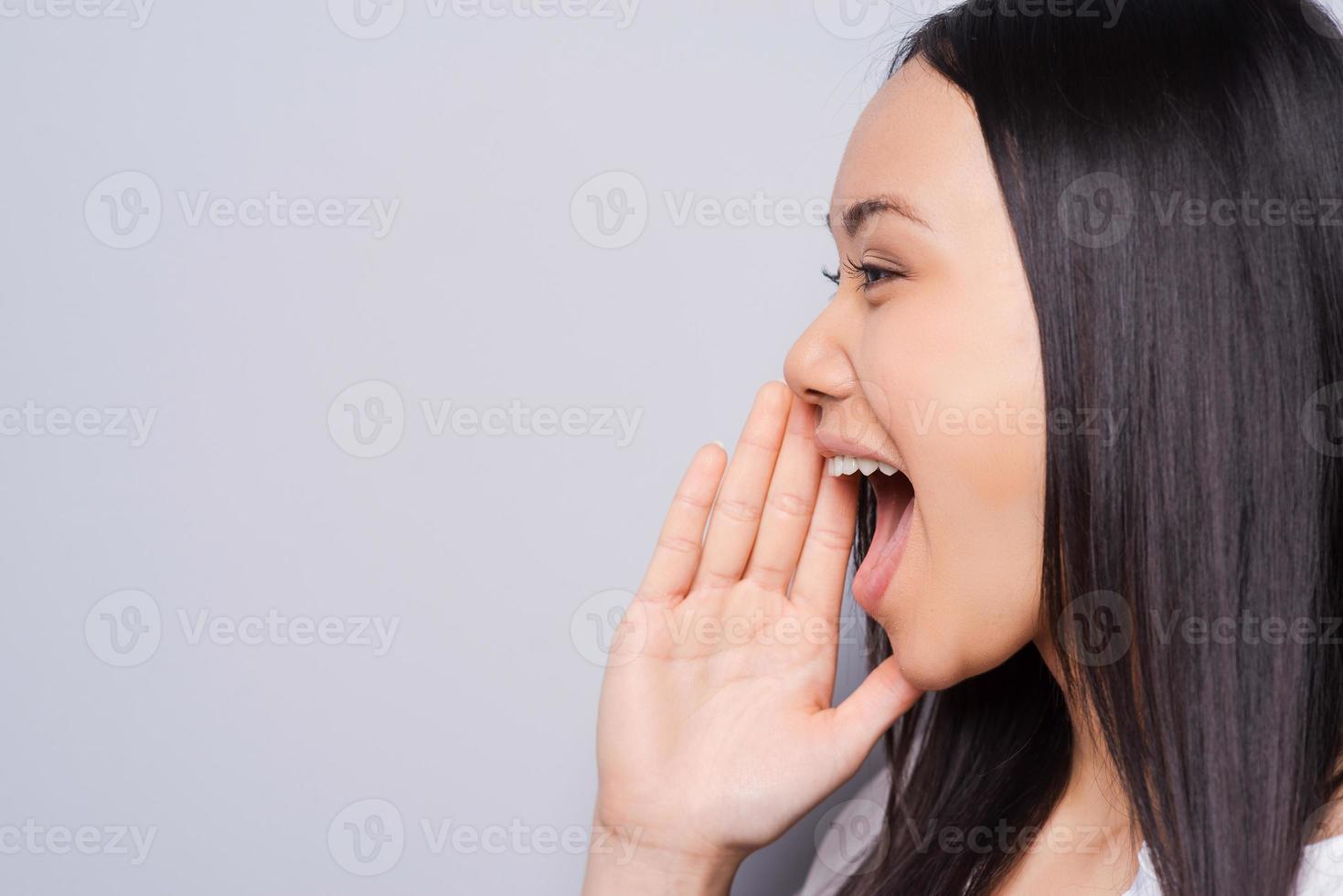 compartiendo buenas noticias. vista lateral de una hermosa joven asiática gritando y sosteniendo la mano cerca de la boca mientras se enfrenta a un fondo gris foto