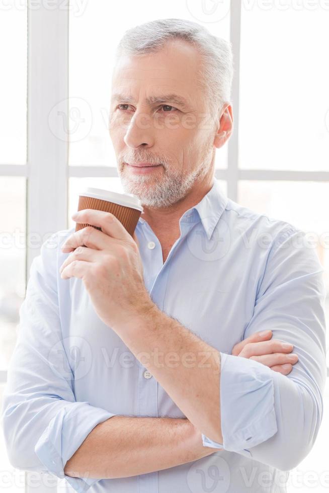 Inspired with cup of fresh coffee. Thoughtful grey hair senior man in shirt holding coffee cup and looking away while standing in front of the window photo