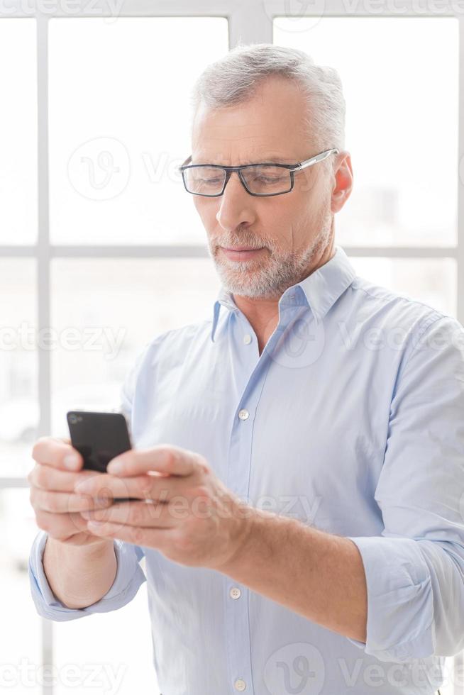 Typing business message. Serious grey hair senior man in shirt holding mobile phone and looking at it while standing in front of the window photo