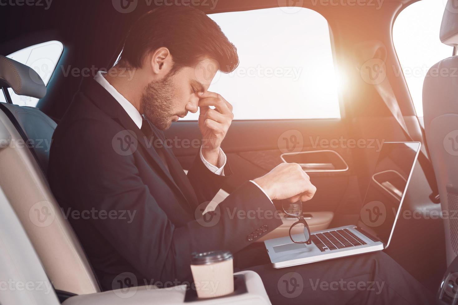 con exceso de trabajo joven frustrado con traje completo masajeando la nariz mientras se sienta en el auto foto