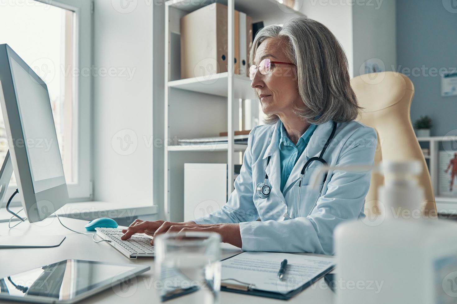 Serious mature female doctor in white lab coat working using computer while sitting in her office photo