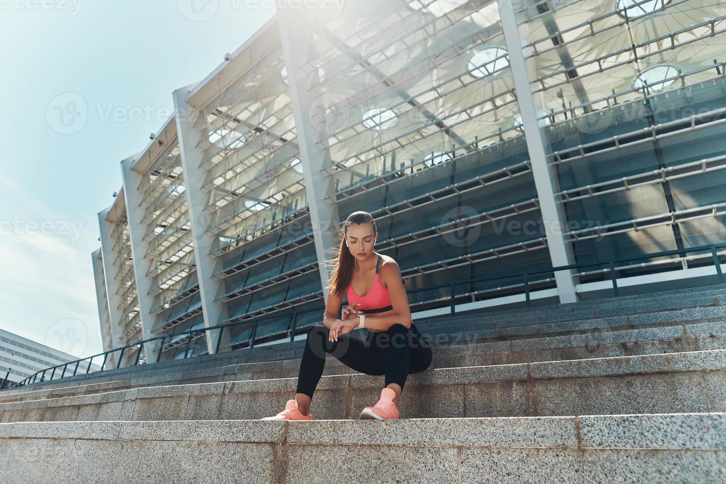 Confident young woman in sportswear checking her smart watch while sitting outdoors photo