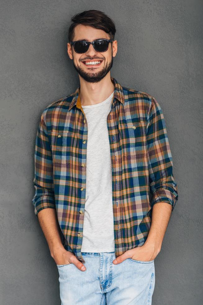 Feeling casual today. Cheerful young man in sunglasses holding hands in pockets and looking at camera with smile while standing against grey background photo