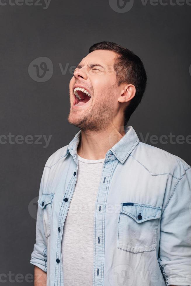 So furious Young handsome man keeping mouth open and eyes closed while standing against grey background photo