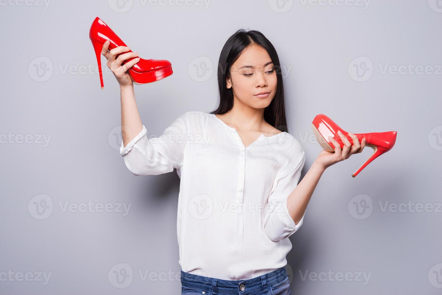 Eternal dilemma. Beautiful young Asian woman holding shoes of different colors while standing against grey background photo