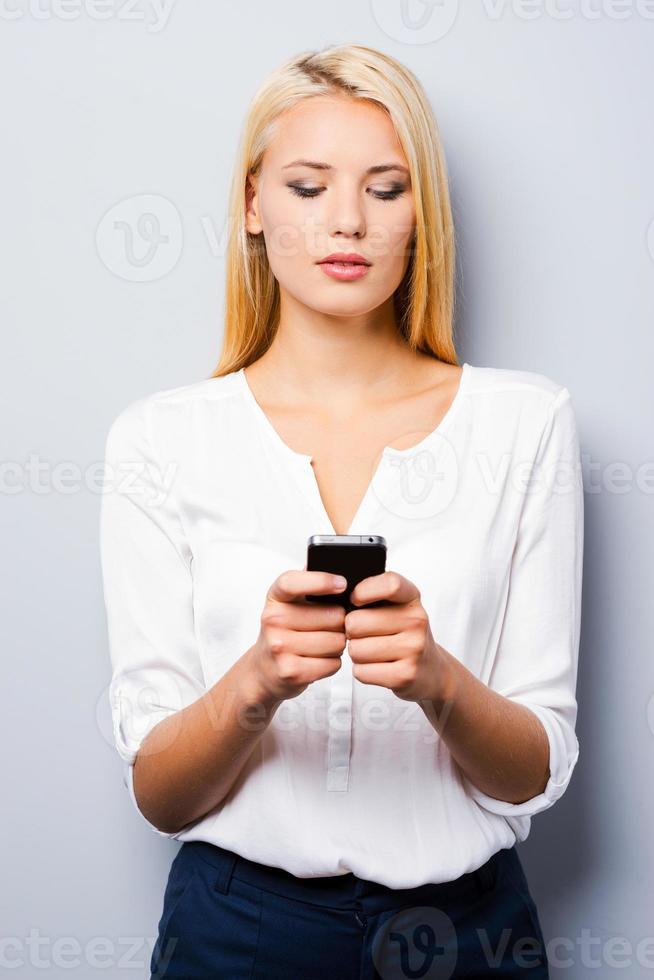 Typing a message for you. Beautiful young blond hair women holding mobile phone and looking at it while standing against grey background photo