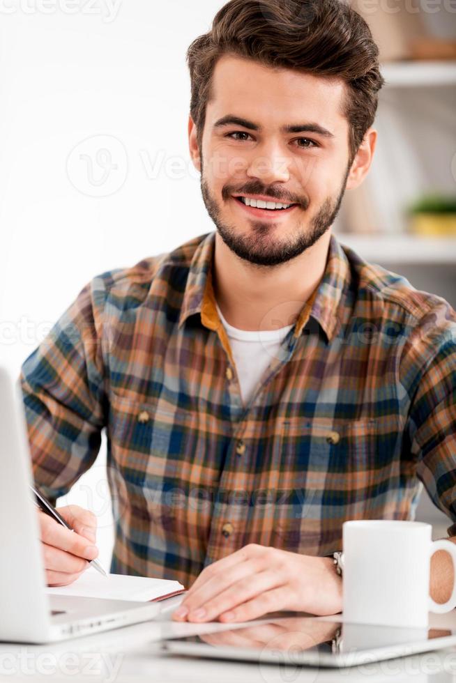 Face of work satisfaction. Smiling young man writing in note pad and looking at camera while sitting at his working place photo