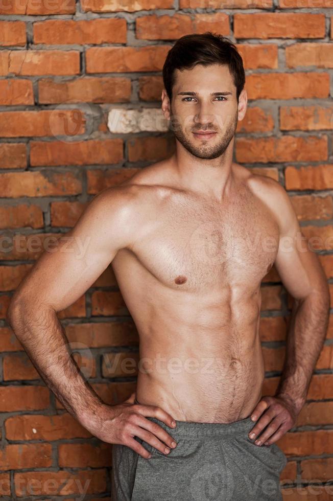 Strong handsome. Handsome young shirtless man holding hands on hips and looking at camera while standing against brick wall photo