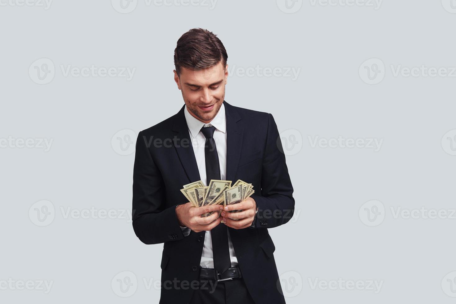 Always in profit. Good looking young man in full suit counting money and smiling while standing against grey background photo