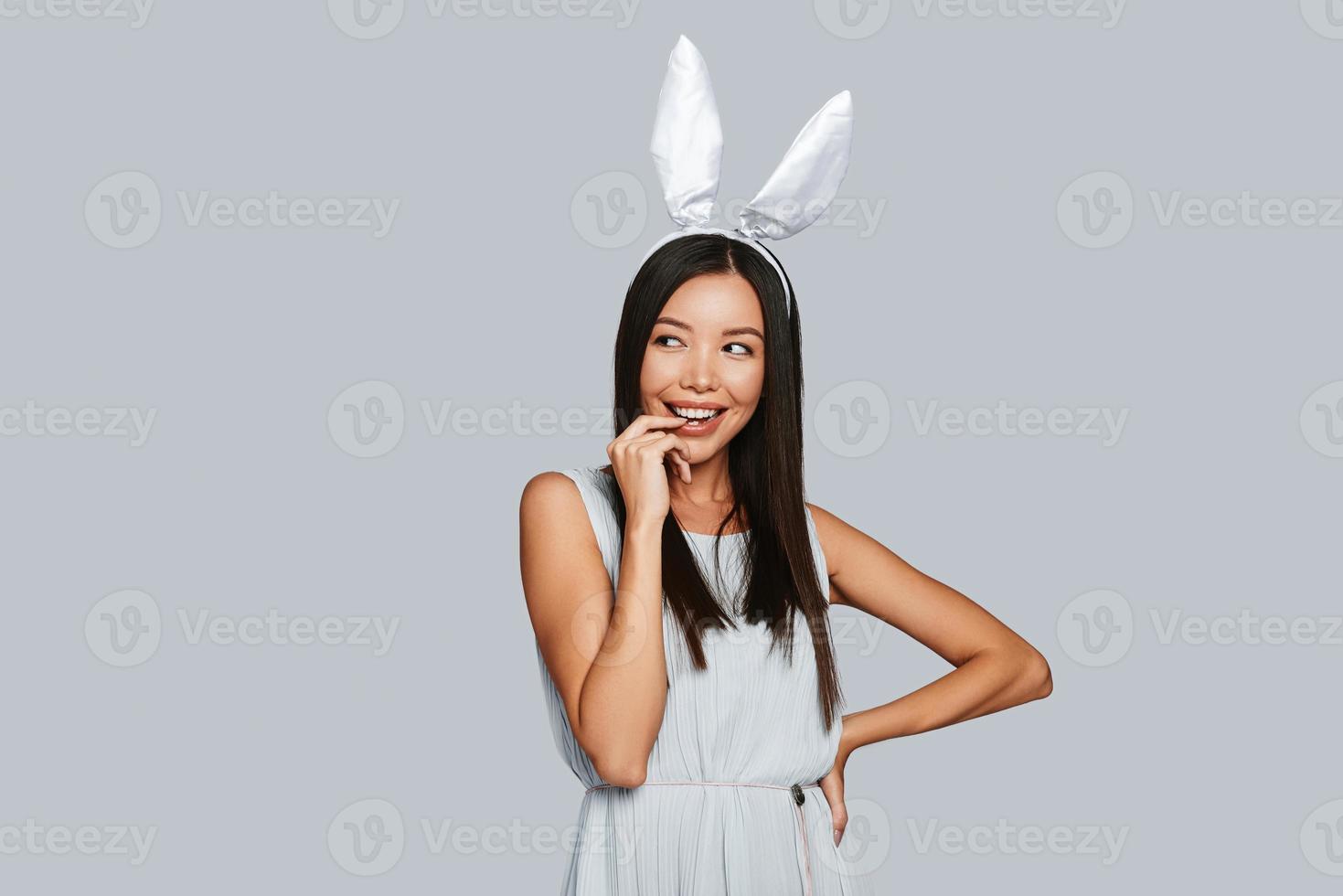 What is there Curios young Asian woman in bunny ears smiling while standing against grey background photo