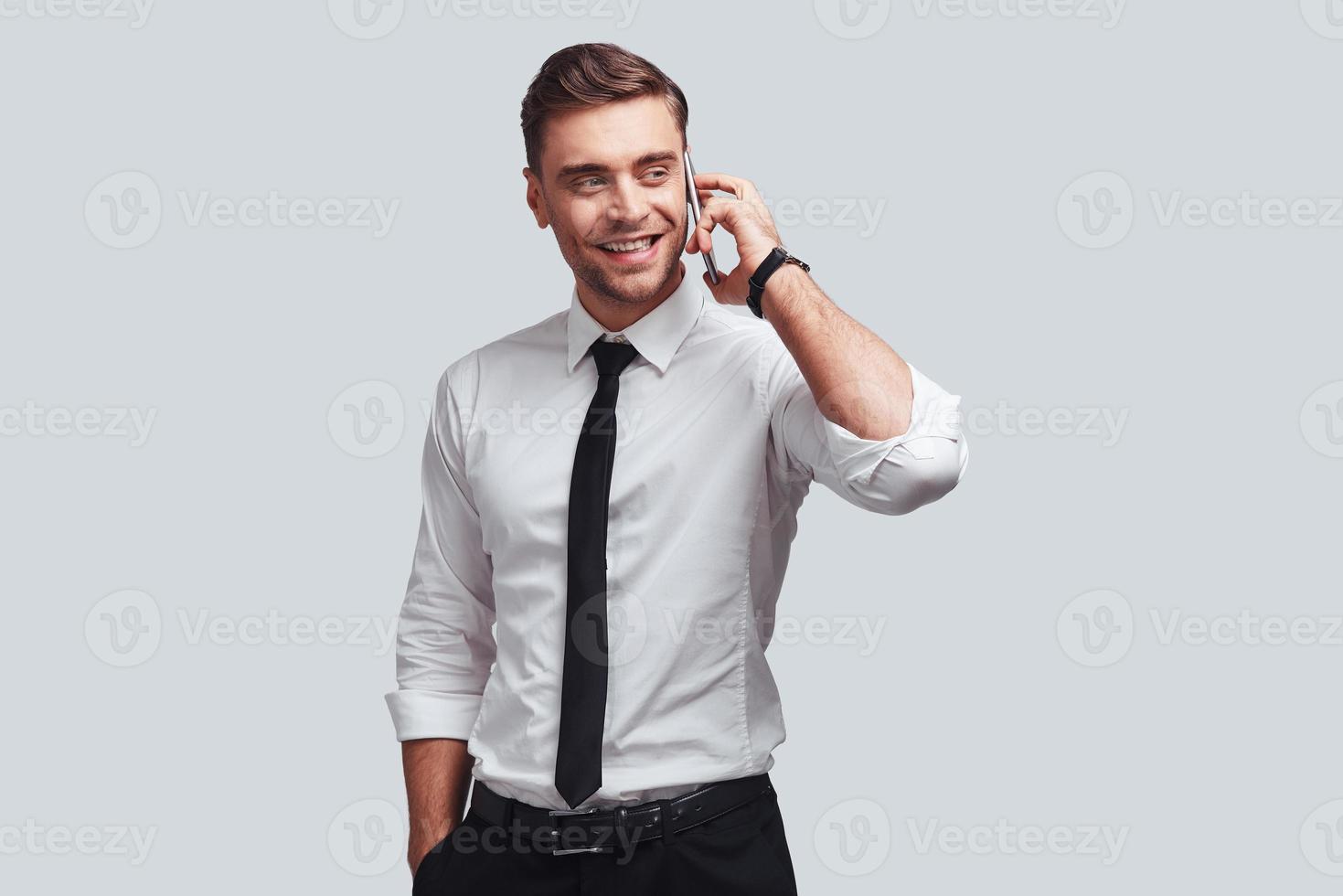Talking with client. Handsome young man in shirt and tie talking on smart phone and looking away with smile while standing against grey background photo
