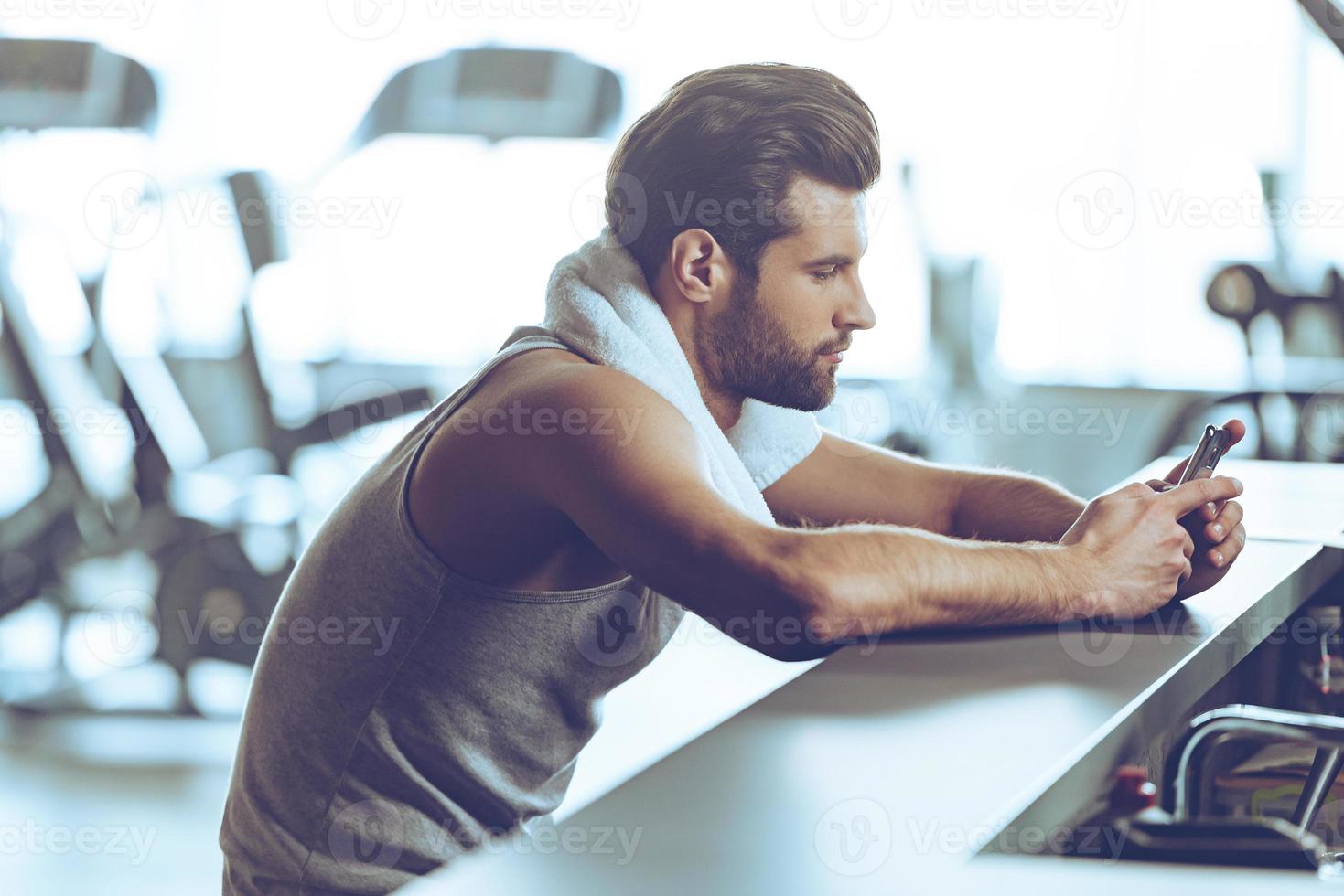 Staying in touch at gym. Side view of handsome young men in sportswear wearing towel on his shoulders and using his smart phone while sitting at bar counter at gym photo