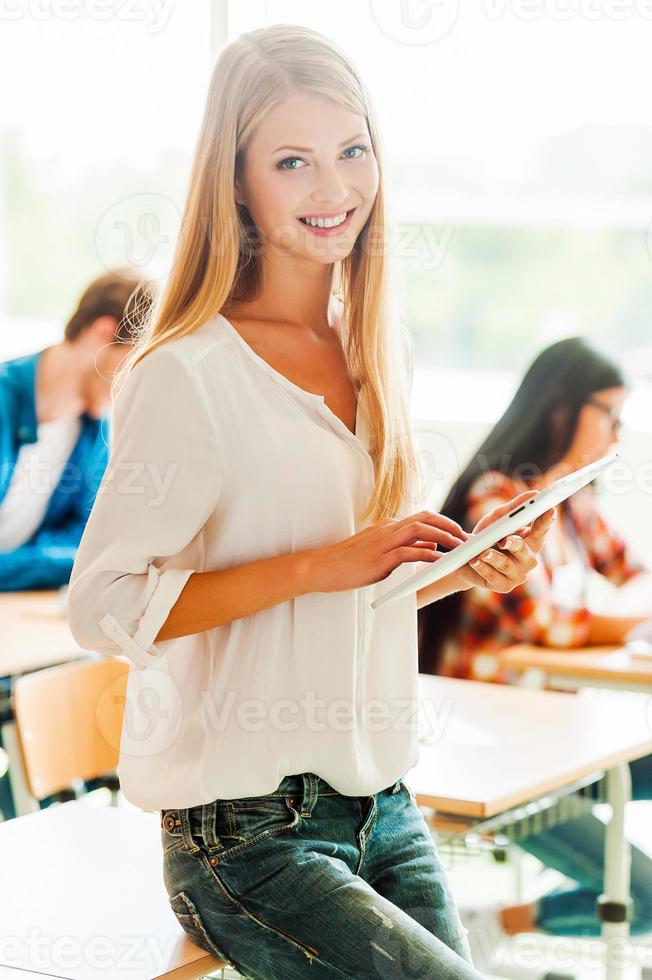 Studying in a digital way. Beautiful young woman holding digital tablet and looking at camera while leaning at the desk photo