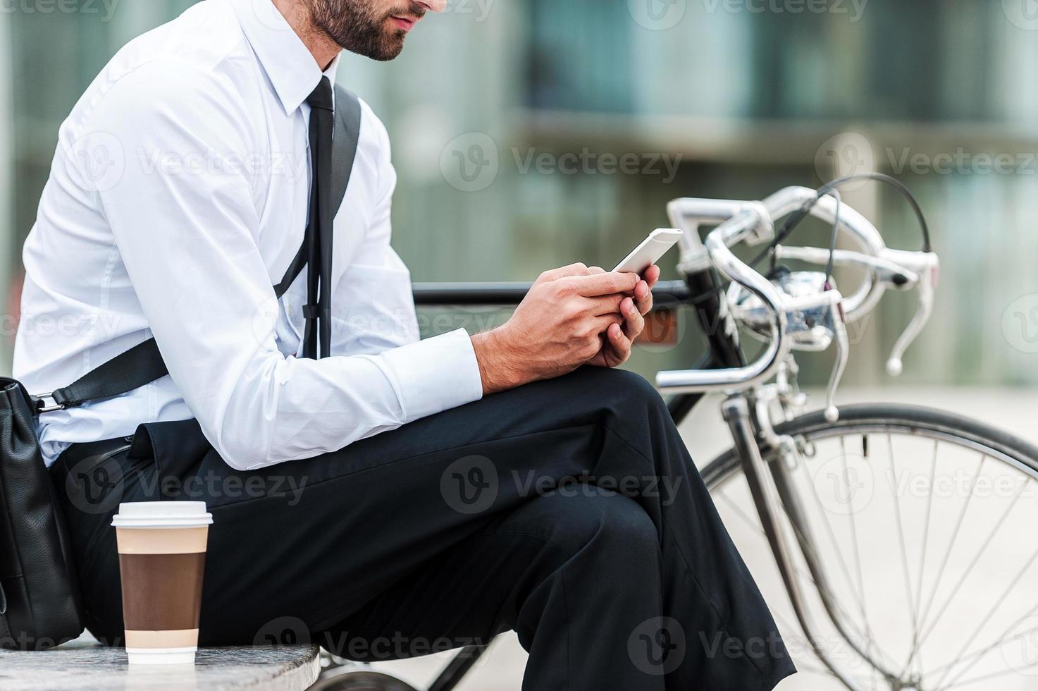 Sending business message. Cropped image of young businessman holding mobile phone while sitting near his bicycle outdoors photo
