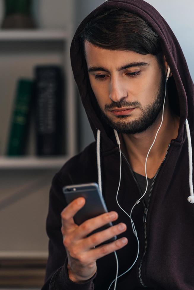 Sad song. Pensive young man in hooded shirt using his smartphone while standing at home photo