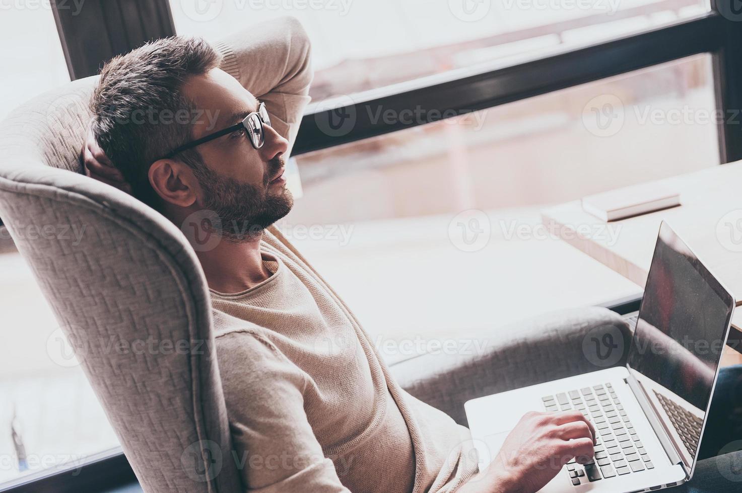 Searching for inspiration. Side view of handsome man using his laptop and looking pensive while sitting in chair in front of window photo
