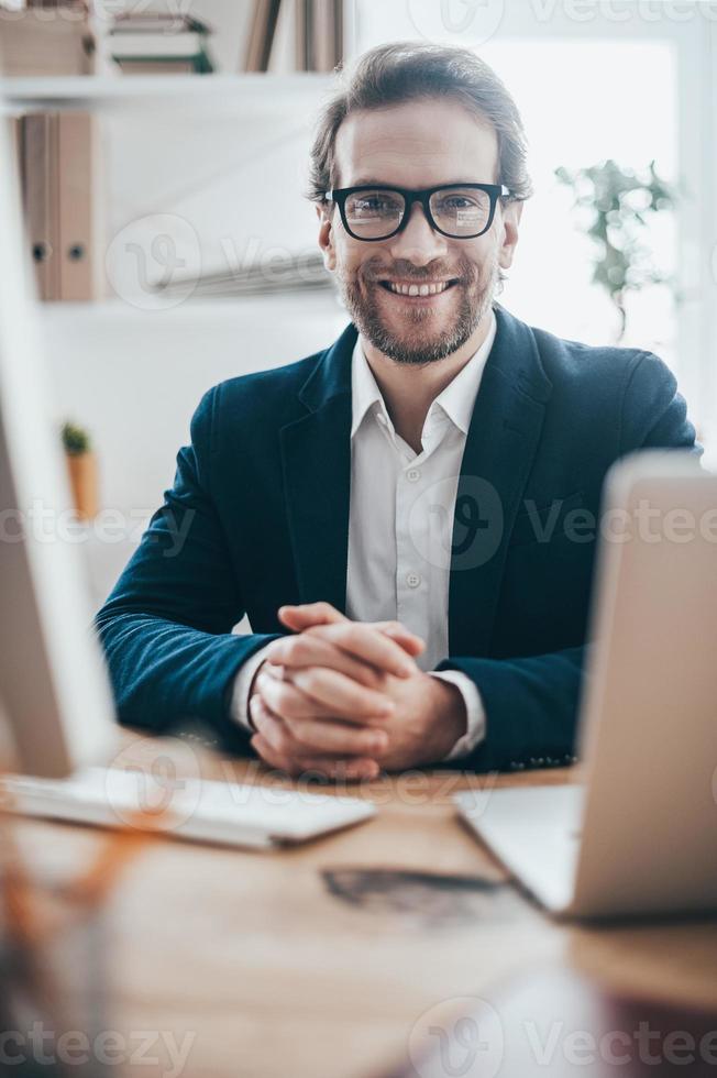 Ready to hear you out. Handsome young man in eyeglasses smiling and looking at camera while sitting on working place in creative office photo