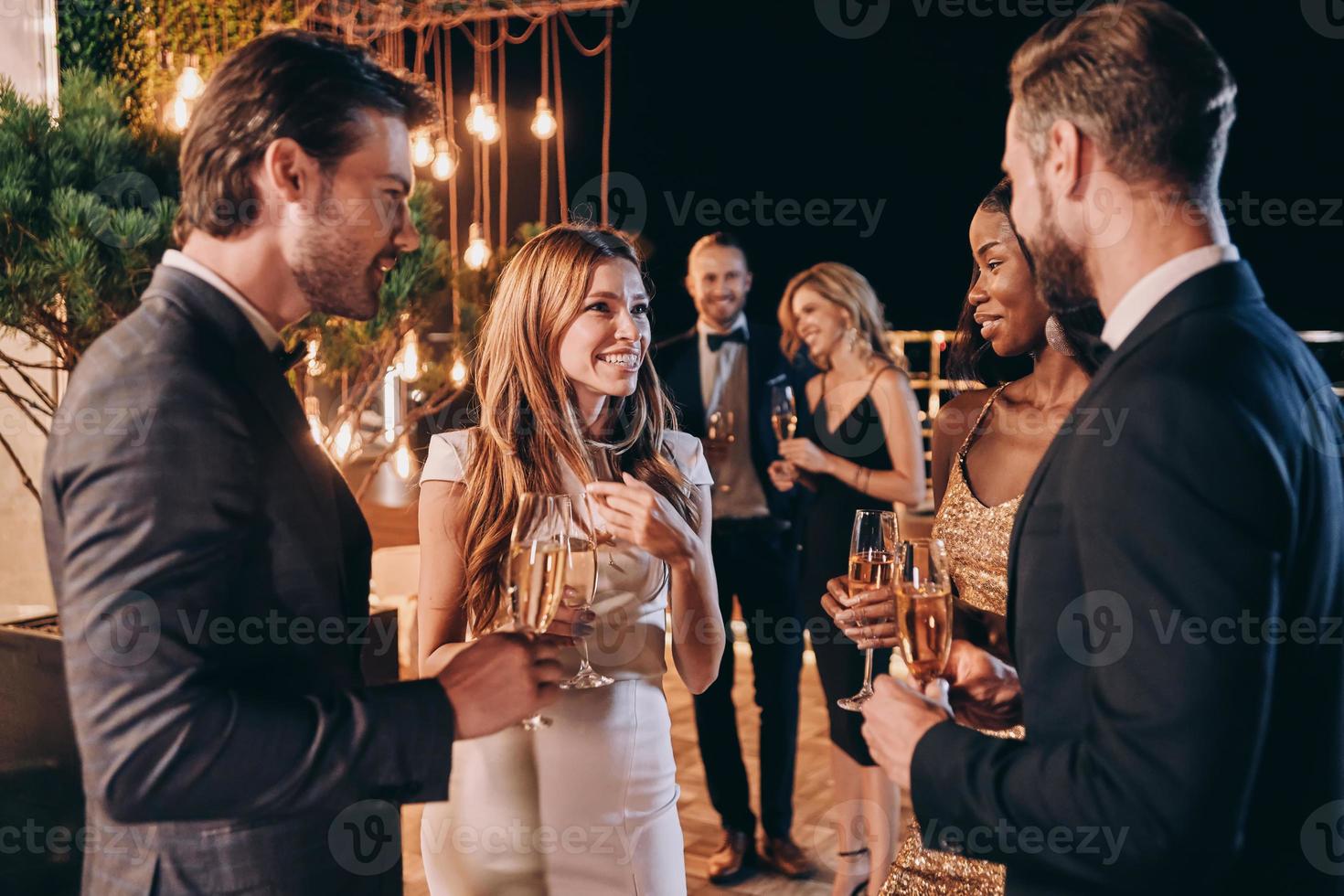 grupo de personas en ropa formal comunicándose y sonriendo mientras pasan tiempo en una fiesta de lujo foto