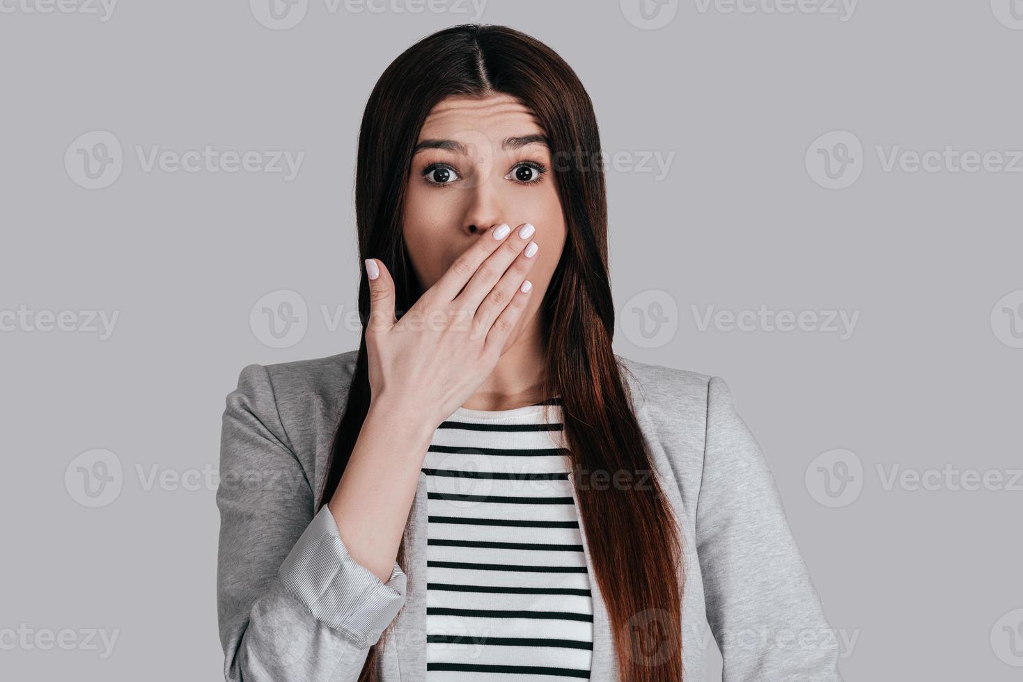 Shocking news Surprised beautiful young woman in smart casual wear covering mouth with hand and looking at camera while standing against grey background photo