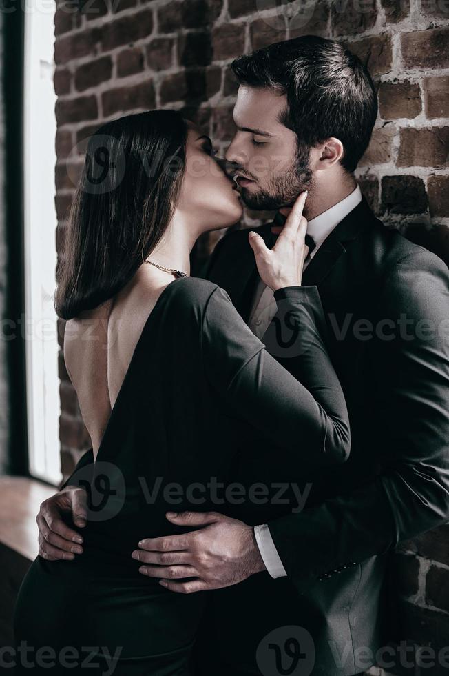 Tender kiss. Beautiful young loving couple bonding to each other and kissing while both standing against brick wall indoors photo