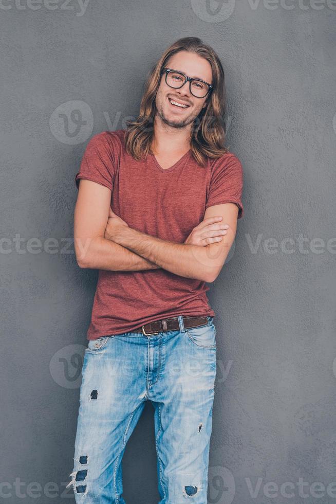 Feeling just casual. Enjoying his casual style. Handsome young man in casual wear and eyeglasses keeping arms crossed and looking at camera with smile while standing against grey background photo