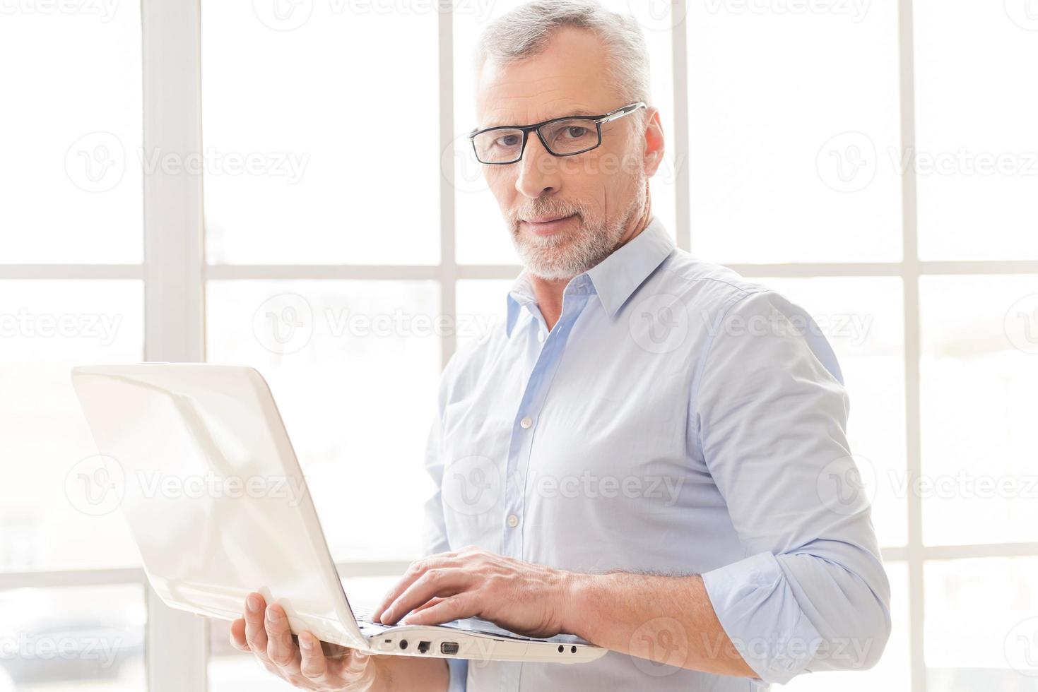 Supporting your business. Confident grey hair senior man in shirt working on laptop and looking at camera while standing in front of the window photo