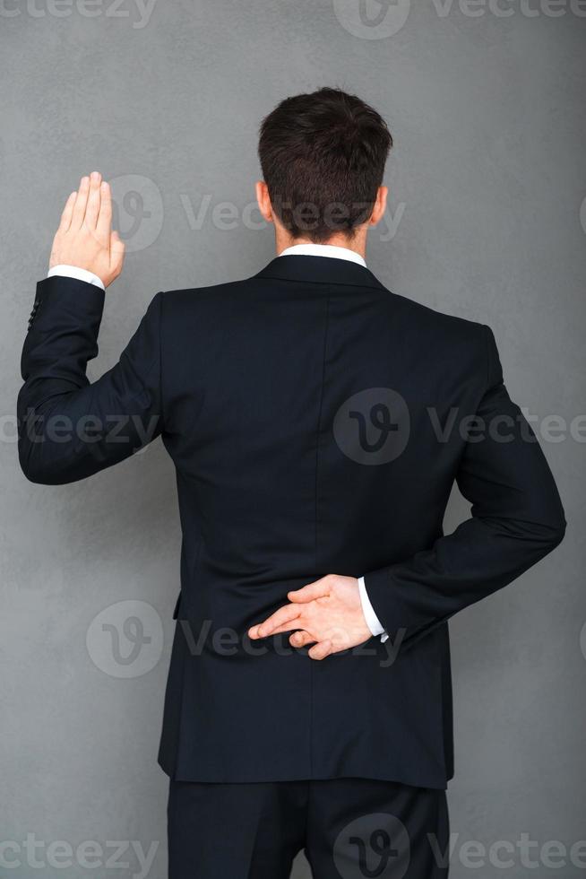 Telling lies. Rear view of young businessman keeping his fingers crossed and arm raised while standing against grey background photo