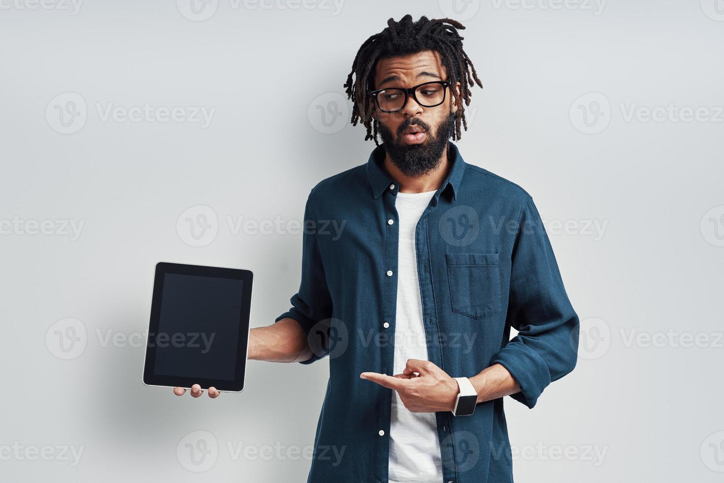 Playful young African man in eyewear pointing copy space using digital tablet while standing against grey background photo