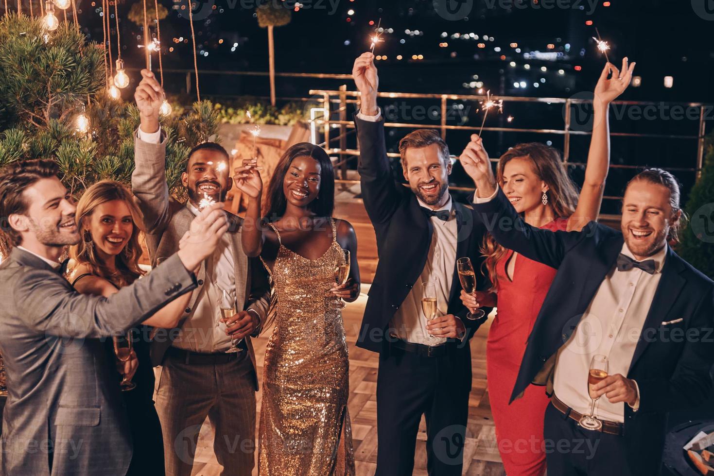 grupo de personas hermosas en ropa formal sosteniendo bengalas y sonriendo mientras pasan tiempo en una fiesta al aire libre foto