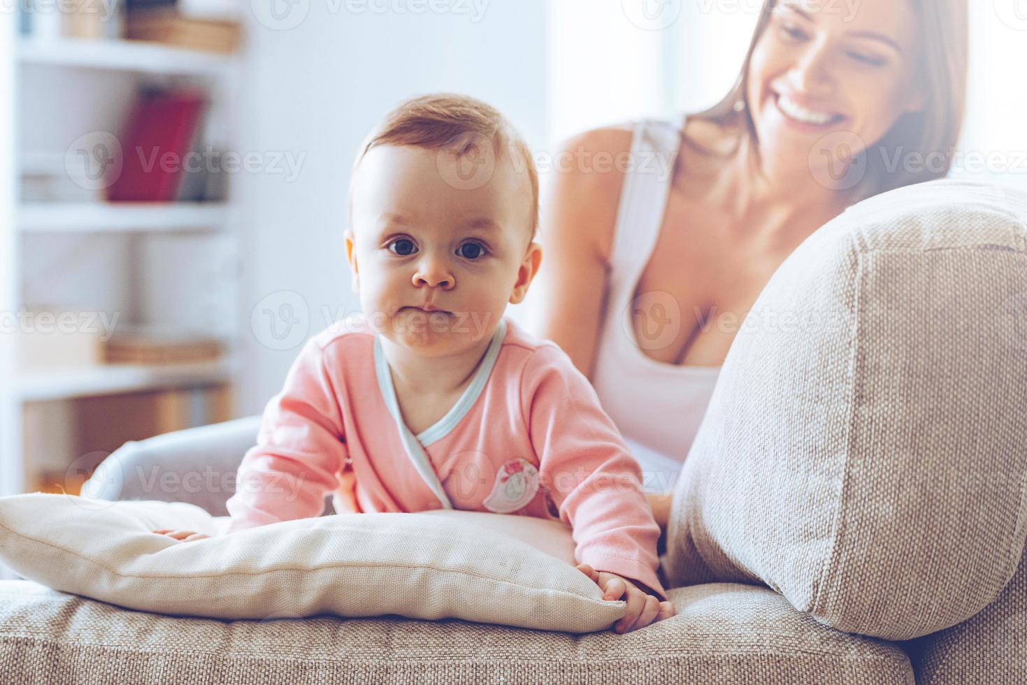 linda con mami. niñita mirando a la cámara y apoyándose en un cojín mientras su madre se sienta en el sofá al fondo foto