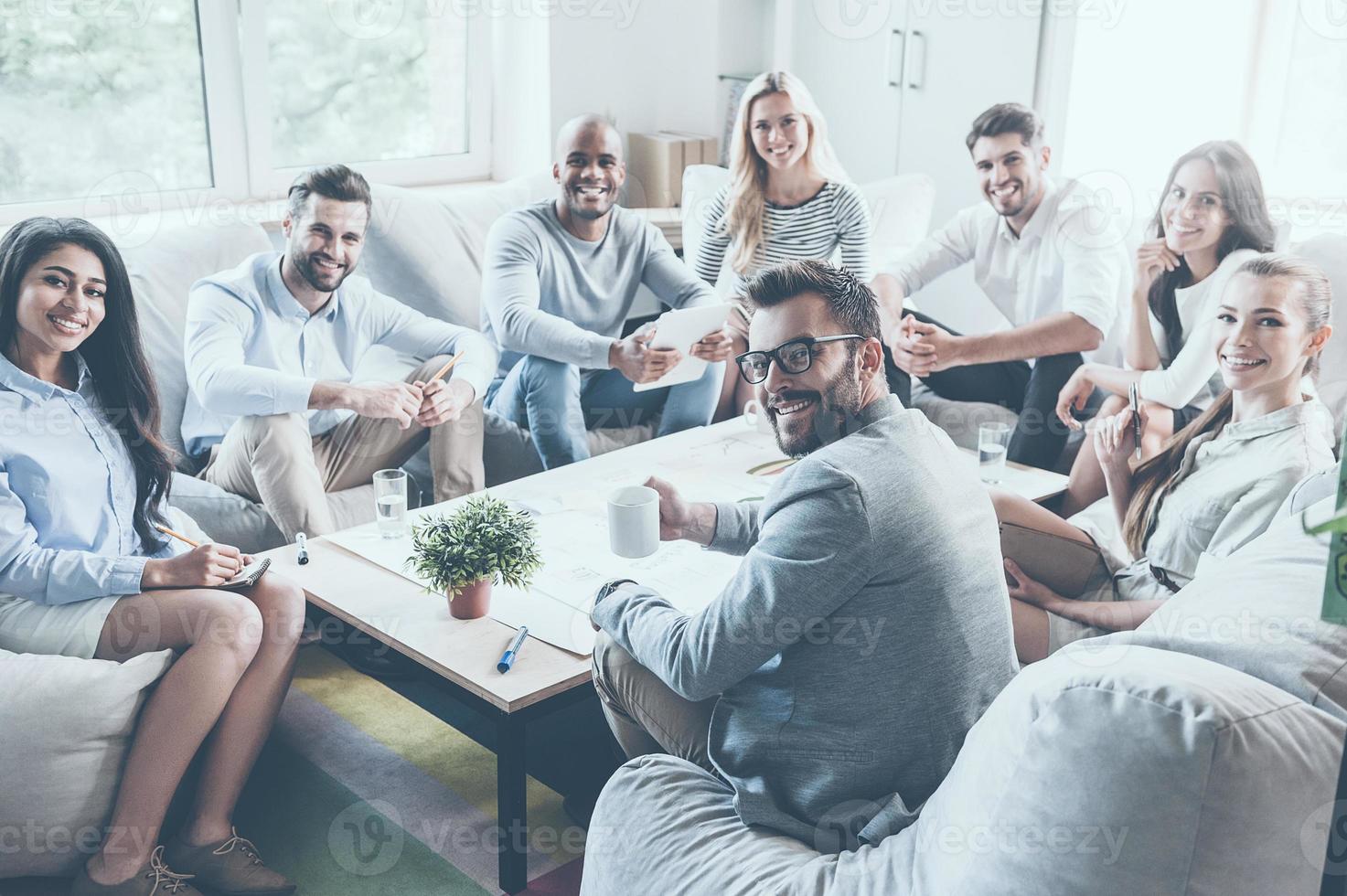 equipo de negocios exitoso. grupo de jóvenes empresarios confiados sentados juntos en el escritorio de la oficina y mirando la cámara con sonrisas foto