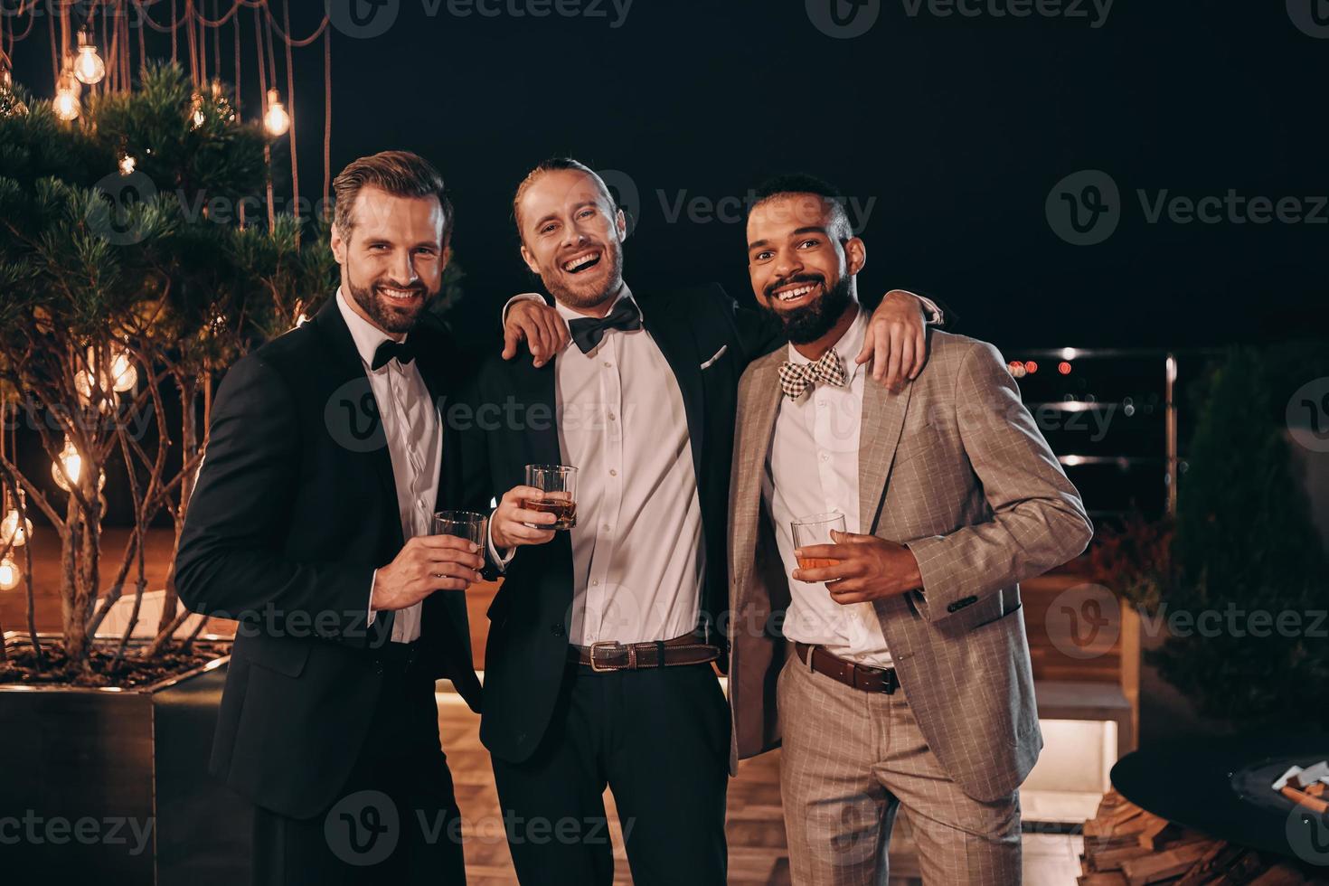 Three cheerful men in suits holding whiskey glasses and looking at camera while spending time on party photo