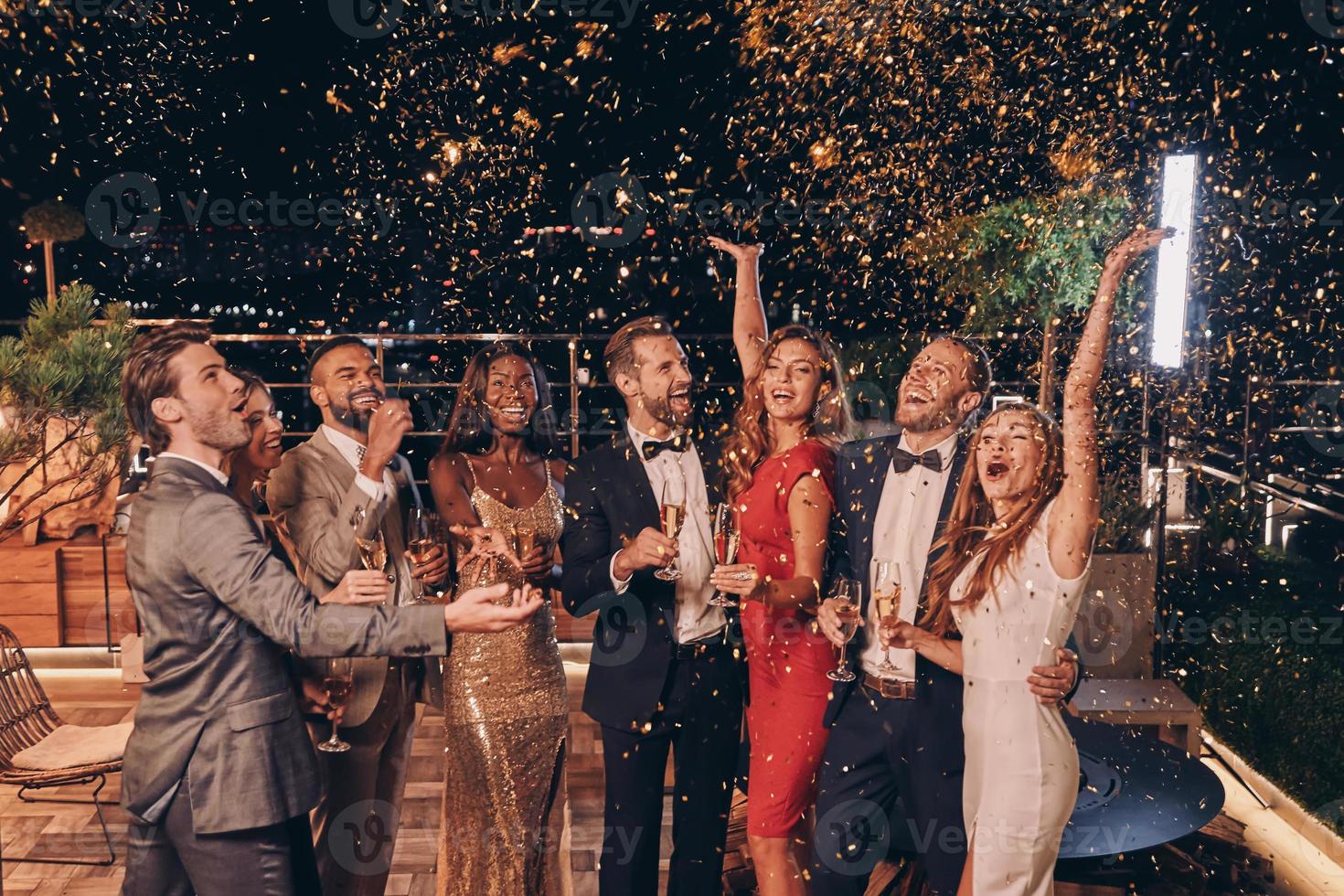 Group of happy people in formalwear having fun together with confetti flying all around photo