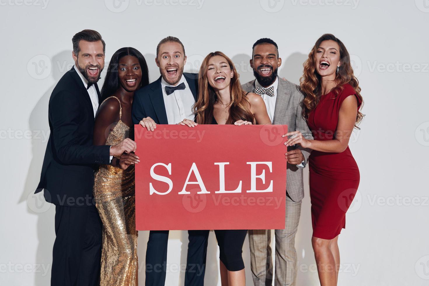 Group of beautiful people in formalwear holding sale banner and smiling while standing against gray background photo