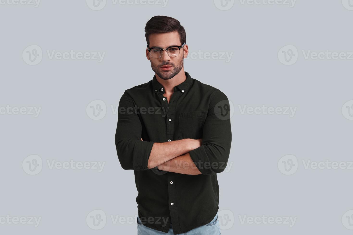 Serious look. Thoughtful young man in eyeglasses keeping arms crossed and looking at camera while standing against grey background photo