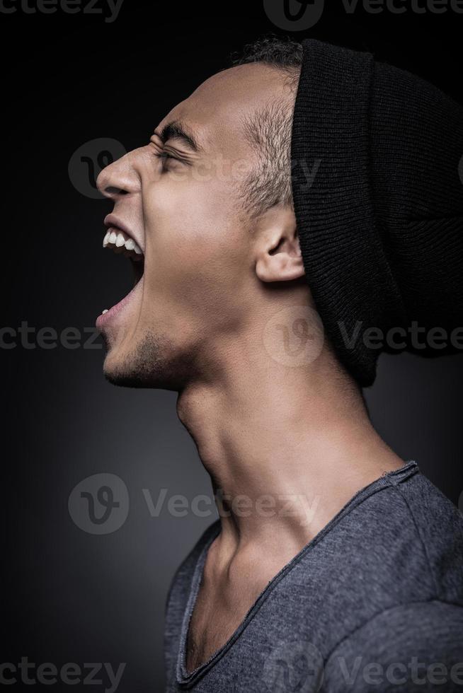 Energy inside him. Side view of furious young African man in beanie keeping eyes closed and mouth open while standing against black background photo