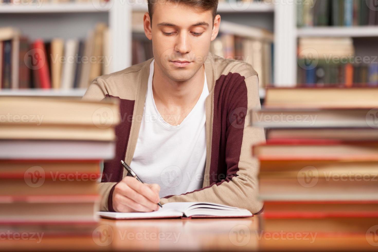 Doing his research in library. Confident young man writing something in his note pad while sitting at the library desk photo