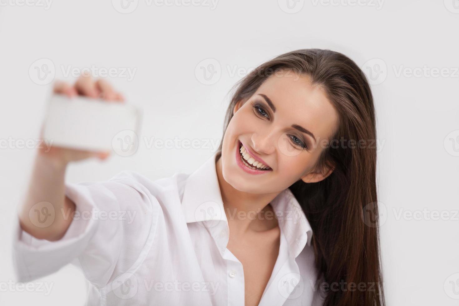 belleza con teléfono móvil. bella joven fotografiando con la cámara de su teléfono móvil y sonriendo mientras está aislada en blanco foto