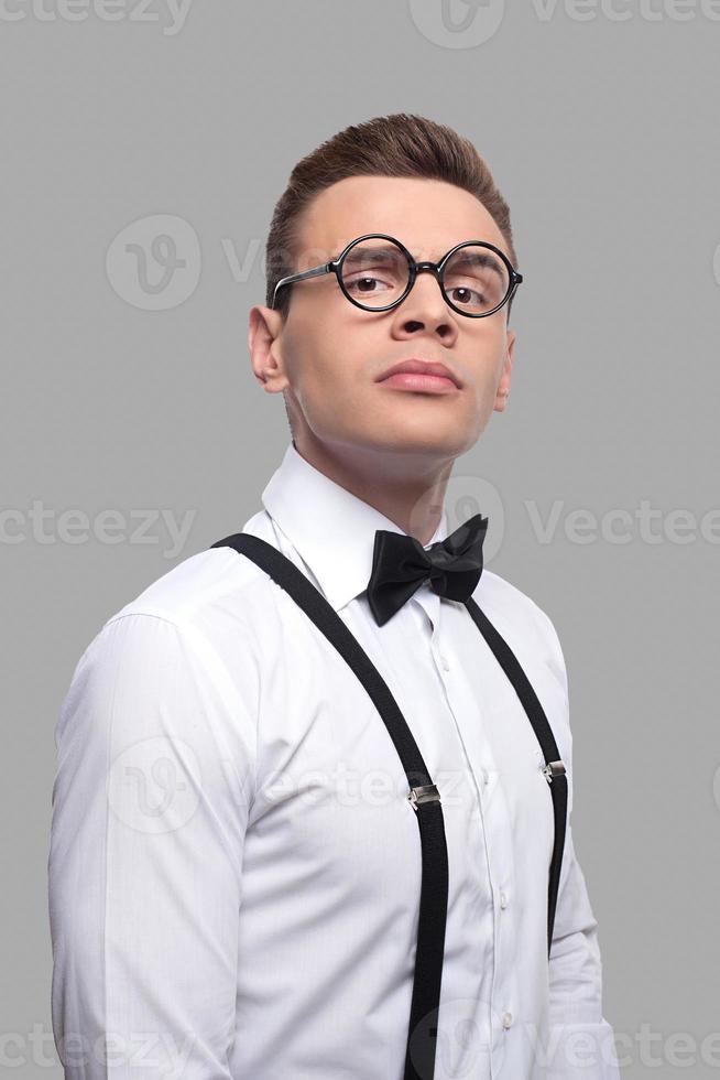 confiado e inteligente. retrato de un joven nerd serio con corbata de moño y tirantes mirando a la cámara mientras se enfrenta a un fondo gris foto