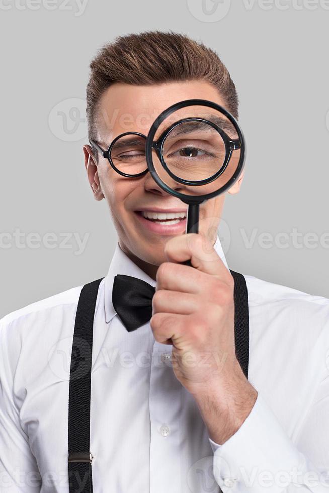 Magnifying you. Portrait of cheerful young man in bow tie and suspenders holding magnifying glass in front of his eye and smiling photo