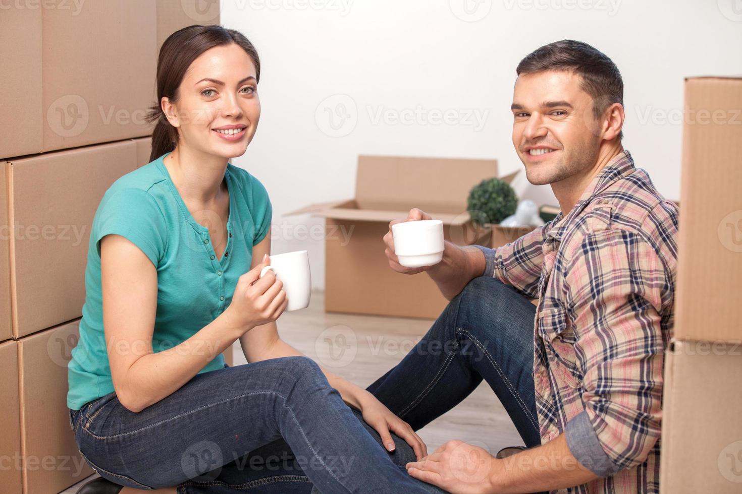Relaxing in new house. Cheerful young couple sitting on the floor and drinking coffee while cardboard boxes laying all around them photo