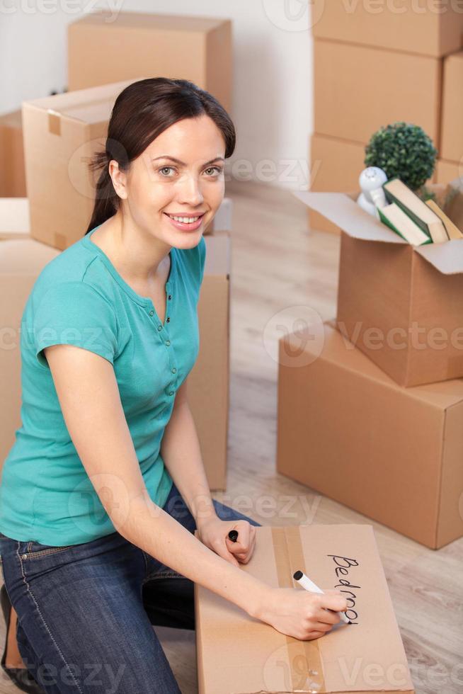 Marking a carton box. Top view of cheerful young woman marking a cardboard box and looking at camera while more boxes laying on background photo