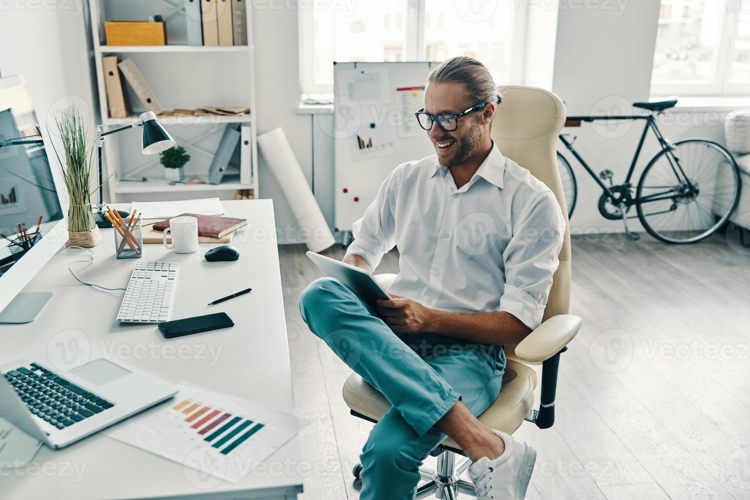 Confident businessman. Top view of good looking young man in shirt using digital tablet and smiling while sitting in the office photo