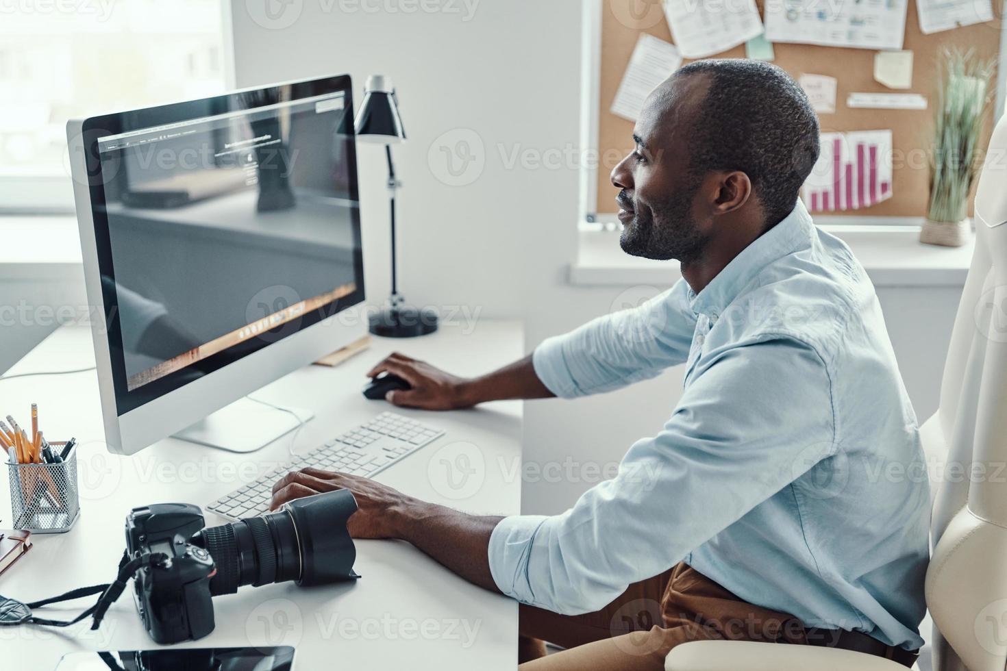 apuesto joven africano usando computadora y sonriendo mientras trabaja en la oficina moderna foto