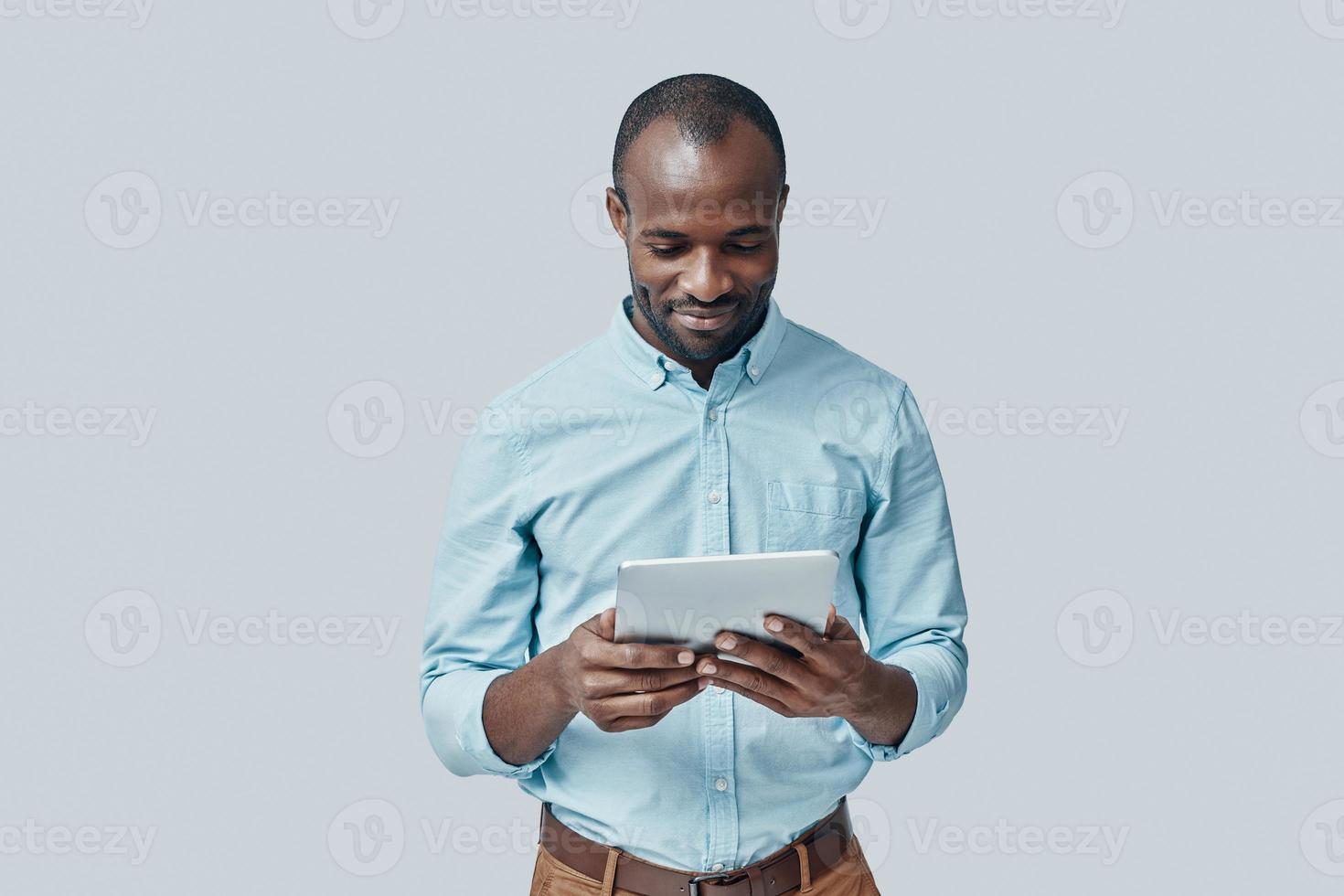Handsome young African man using digital tablet and smiling while standing against grey background photo
