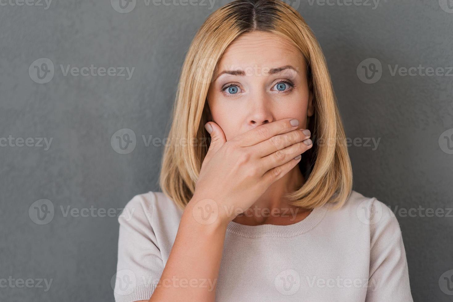 Shocking news. Surprised mature woman covering mouth with hand and staring at camera while standing against grey background photo