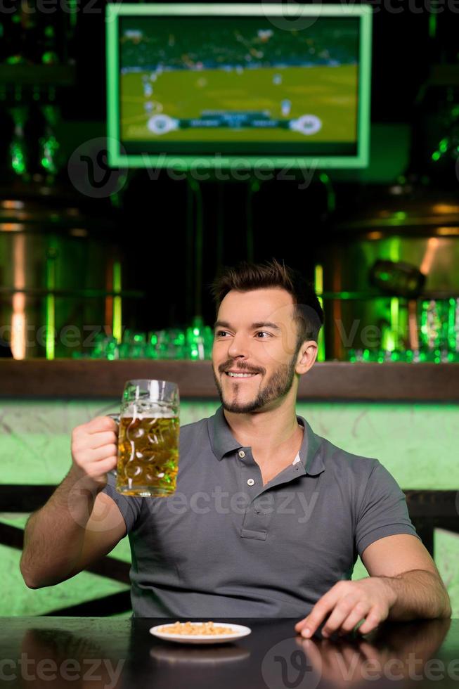Man in beer pub. Cheerful young man holding a mug with beer and smiling while sitting in bar photo