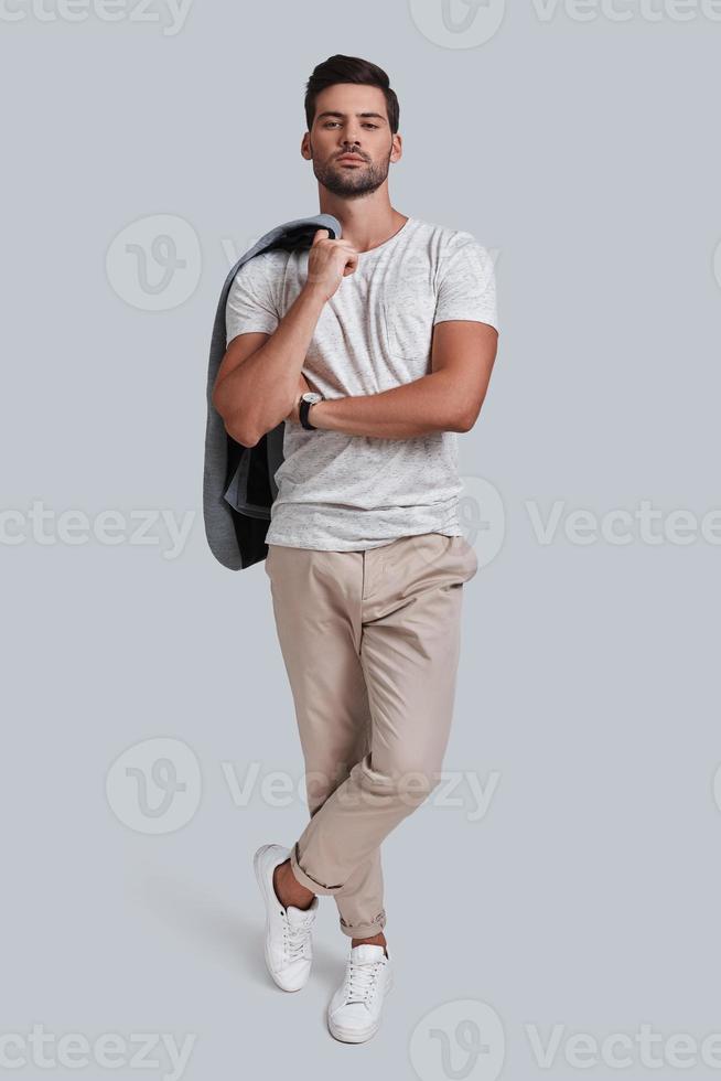 Full length of handsome young man carrying jacket on shoulder and looking at camera while standing against grey background photo