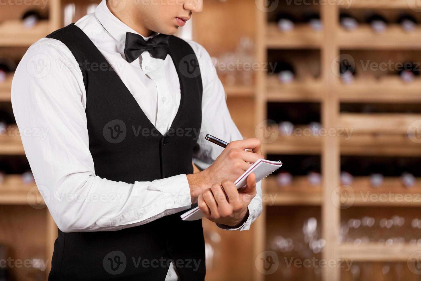 Checking wine list. Cropped image of young sommelier writing something at his note pad while standing in front of shelf with wine bottles photo
