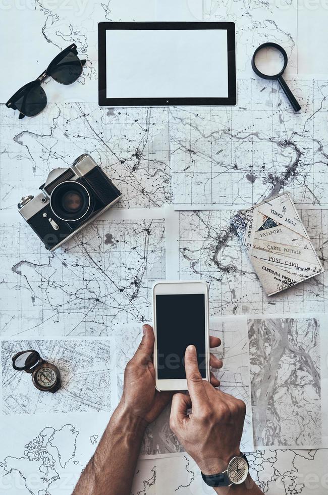 New directions. Close up top view of man using smart phone with sunglasses, photo camera, compass, magnifying glass and passport lying on map around